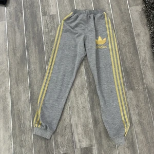 Grå mjukisbyxor från Adidas - Snygga grå mjukisbyxor från Adidas med gula ränder längs sidorna och det klassiska Adidas-loggan på ena benet. Byxorna har elastisk midja och muddar vid bensluten för en bekväm passform.