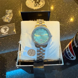 Silverfärgad klocka från Versus Versace - Funkar som nytt.  Elegant silverfärgad klocka från Versus Versace med en skimrande urtavla och dekorativa stenar. Klockan har en robust länkarmband och ett lejonhuvud som detalj på urtavlan. Perfekt för den som vill ha en stilren accessoar med en touch av lyx. 