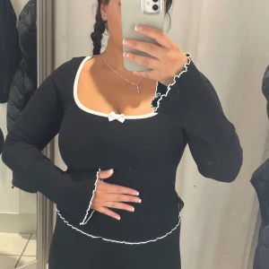 Svart ribbad topp med vita detaljer - Snygg svart ribbad topp med långa ärmar och vita kanter. Toppen har en söt rosett framtill och en v-ringad design. Perfekt för en stilren look.