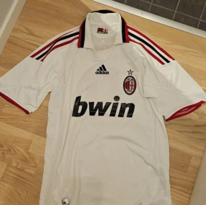Retro AC Milan fotbollströja från säsongen 2009/2010 - Säljer en retro AC Milan fotbollströja från Adidas med korta ärmar och röda detaljer. Tröjan har klubbens emblem och sponsorlogga på framsidan samt texten 'Forza Milan' på insidan av kragen. Perfekt för fans av laget!