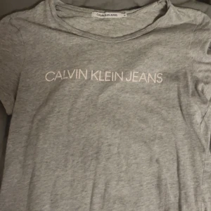 Grå t-shirt från Calvin Klein Jeans - Säljer en stilren grå t-shirt från Calvin Klein Jeans med korta ärmar. T-shirten har en klassisk passform och är tillverkad i mjukt material med märkeslogga framtill. Perfekt för en avslappnad look.