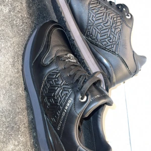 Svarta sneakers från Tommy Hilfiger - Snygga svarta sneakers från Tommy Hilfiger med präglat mönster på sidorna och logotypdetalj på snörningen. Storlek 40, knappt använda! 