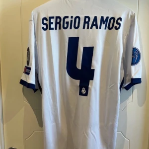 Vit Real Madrid fotbollströja med Sergio Ramos  - Vit Real Madrid fotbollströja från Adidas med Sergio Ramos namn och nummer 4 på ryggen. Tröjan har korta ärmar och klubbens emblem på bröstet. Den har även FIFA-märket och Champions League-loggor på ärmarna.