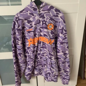 Lila camo hoodie från Bape - Säljer en cool lila camo hoodie som liknar Bape med dragkedja och huva. Den har ett iögonfallande orange tryck på framsidan och är perfekt för streetwear-stilen. Hoodie är långärmad och har en avslappnad passform. Köpt i tyskland och helt oanvänd! Pris kan diskuteras😊