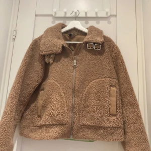 Beige teddyjacka  - Mysig beige teddyjacka från H&M med dragkedja och stora fickor framtill. Jackan har en hög krage med spännen och är perfekt för kyligare dagar. Den har en avslappnad passform och är superbekväm att bära.