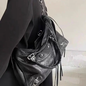 Svart axelväska/ handväska - Snygg svart axelväska i skinn med silverfärgade metalldetaljer. Väskan har en justerbar axelrem och flera dragkedjor för praktisk förvaring. Perfekt för att ge din outfit en edgy touch. Väskan är aldrig använd.