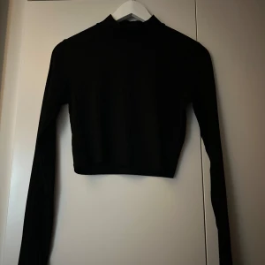 Svart ribbad croppad topp - Snygg svart ribbad topp med lång ärm och croppad passform. Perfekt för en stilren look. Toppen har en hög krage som ger en elegant touch. Passar inte mig längre därav säljer jag