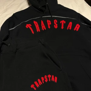 Trapstar tracksuit - Snygg svart tracksuit från Trapstar med röd text  Den har en klassisk design med huva och dragsko. Perfekt för en avslappnad stil.