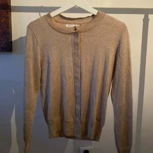 Glittrig beige kofta från Holly & Whyte - Snygg beige kofta från Holly & Whyte med glittrande effekt. Den har långa ärmar och knappar framtill. Perfekt för att lägga till lite glamour i vardagen. ✨