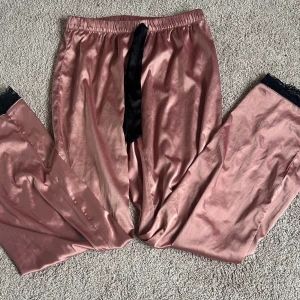 Rosa satinbyxor med spetsdetaljer - Snygga rosa byxor i satin med elastisk midja och svart knytband. Byxorna har spetsdetaljer vid bensluten som ger en elegant touch. Perfekta som pyjamasbyxor 