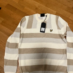 Randig tröja från Lyle & Scott - Säljer en snygg randig tröja från Lyle & Scott i off-white och beige. Tröjan har ett broderat emblem på bröstet och är perfekt för en stilren look. Den är långärmad och tillverkad i ett bekvämt material.