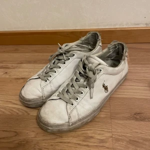 Vita sneakers från Polo Ralph Lauren - Säljer ett par vita sneakers från Polo Ralph Lauren med klassisk design och logotyp på sidan. Skorna har snörning och är tillverkade i skinn. Perfekta för en avslappnad stil. De är slitna där bak men man tänker inte på det. 
