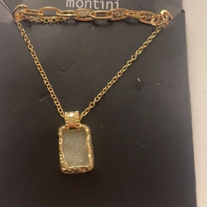 Guldhalsband med rektangulär hängsmycke från Montini - Elegant guldhalsband från Montini med en rektangulär hängsmycke. Hängsmycket har en glittrig yta och är inramat i en dekorativ guldfärgad kant. Kedjan är delikat och kompletterar det stilrena utseendet.