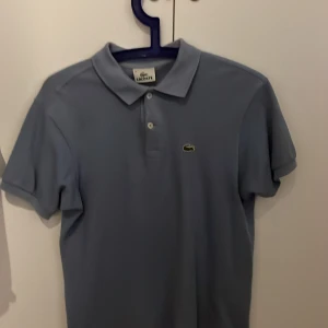Ljusblå pikétröja från Lacoste - Det är storlek 14. Alltså de passar personer runt 14 år. Jag skulle säga att den passar om man är mellan 160-175. Snygg ljusblå pikétröja från Lacoste med klassisk krage och knappar framtill. Tröjan har det ikoniska krokodilmärket på bröstet. Inga defekter eller slitningar.                                Hör av er vid frågor eller funderingar! Mvh Melker
