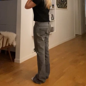 Grå jeans med slitningar - Säljer ett par grå jeans med slitningar på knäna. De har en bootcut-stil och är lågmidjade. Perfekta för en avslappnad look.