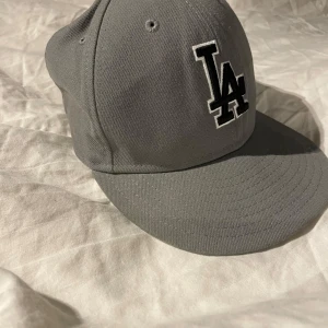 Grå keps från New Era - Snygg grå keps från New Era med LA-logga framtill. Kepsen har en klassisk design med böjd skärm och är tillverkad i bomull. Perfekt för en casual look. Storlek står i sista bilden 