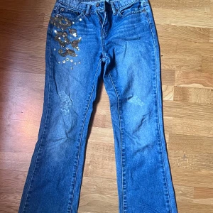 Blå jeans med paljettdetaljer från DKNY - Snygga blå jeans från DKNY med unika guldiga paljettdetaljer på framsidan och bakfickorna. Ganska lågmidjade och lite flare.