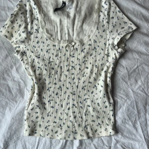 Vit blommig topp från H&M - Söt vit topp från H&M med små blå blommor och ribbad struktur. Den har korta ärmar och spetsdetaljer vid halsringningen samt en liten rosett framtill. Passar xs-s🫶 använd ett fåtal gånger men inget som märks på den