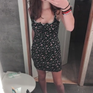 Blommig kortklänning - Söt blommig kortklänning med smala axelband och en figurnära passform. Perfekt för sommarens varma dagar. Klänningen har ett charmigt blommönster i olika färger mot en mörk bakgrund.