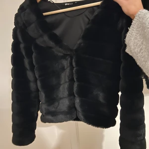 Svart pälsjacka från Gina Tricot - Säljer en elegant svart pälsjacka från Gina Tricot. Jackan har en mjuk och lyxig känsla med långa ärmar och en stilren design. Perfekt för att hålla sig varm och snygg under kyliga dagar.