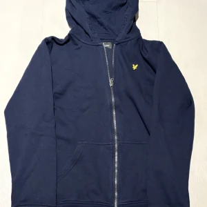 Mörkblå hoodie från Lyle & Scott - Säljer en snygg mörkblå hoodie från Lyle & Scott med dragkedja och gul logga på bröstet. Perfekt för en avslappnad stil. Passar ungdomar i åldern 12-13 år och är ish xs. Pris kan diskuteras vid snabb affär!