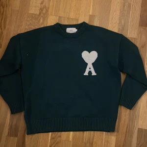 Ami paris stickad - Säljer en snygg mörkgrön stickad tröja från Ami med ett vitt hjärtmönster och bokstaven A på bröstet. Tröjan har långa ärmar och ribbade kanter vid hals, ärmslut och nederkant. Perfekt för kyliga dagar!