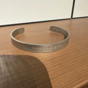 Silverfärgat armband från Maison Margiela - Elegant och stilrent armband i matt silverfärg från Maison Margiela. Armbandet har en minimalistisk design med ingraverad text på framsidan. Perfekt för att ge en sofistikerad touch till din stil.