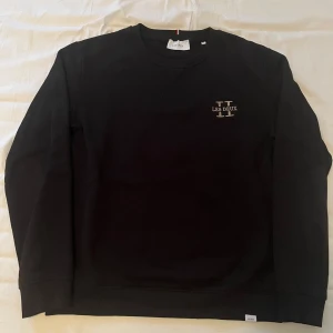 Svart sweatshirt från Les Deux - Snygg svart sweatshirt från Les Deux med deras logga på bröstet. Tröjan har långa ärmar och en rund halsringning med ribbade kanter. Den är i super fint skick helt utan skador och defekter