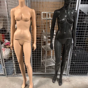 Två skyltdockor i olika färger - Två skyltdockor, en i beige och en i svart, står bredvid varandra. De är helkroppsmodeller utan kläder, placerade i en industriell miljö med metallgaller i bakgrunden.