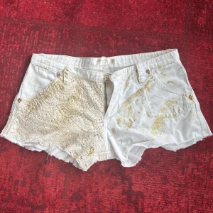 Vita shorts med spets och guldglitter - Unika vita shorts med en mix av spets och denim. Ena sidan är täckt av vit spets medan den andra har guldglitterdetaljer. Perfekta för en somrig look med en twist. Knapparna har en cool design som ger extra stilpoäng.