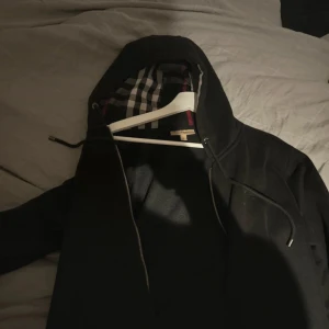 Svart tröja med huva från Burberry - Säljer en stilren svart jacka  från Burberry med klassiskt rutigt foder i huvan. Jackan har dragkedja och en modern design som passar perfekt för höst och vår. Perfekt för dig som vill ha något snyggt och tidlöst i garderoben.