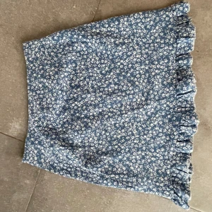 Blå blommig kjol med volang - Söt blå kjol med vita blommor och volangdetalj längst ner. Perfekt för en somrig look. Kjolen har en kort längd och ger en lätt och luftig känsla.