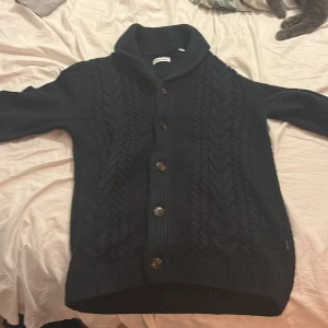 Svart stickad kofta med knappar - Säljer en svart stickad kofta med snygga knappar framtill. Den har en hög krage och långa ärmar, perfekt för kyliga dagar. Koftan har ett klassiskt flätmönster som ger en stilren look.
