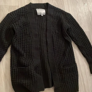 Svart stickad kofta från 157 - Säljer en svart stickad kofta från 157. Koftan har en grovstickad textur och långa ärmar. Perfekt för att hålla sig varm under kyliga dagar. Den har en öppen design utan knappar och ribbade kanter.