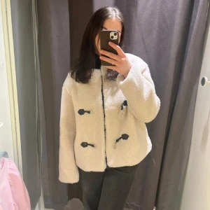 Vit teddyjacka med knäppning - Mysig slutsåld vit teddyjacka från Gina med unik knäppning framtill. Nästan oanvänd. Jackan har en rund hals och långa ärmar, perfekt för kyligare dagar. Den fluffiga texturen ger en trendig look. 