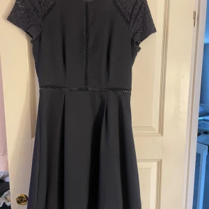 Mörkblå klänning från New Eve - Elegant mörkblå klänning från New Eve med spetsdetaljer på axlarna och korta ärmar. Klänningen har ett fint mönster och en diskret dragkedja i ryggen. Perfekt för en stilren look.