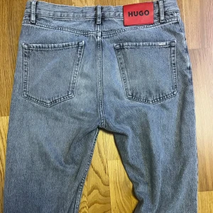 Hugo Boss Jeans - Säljer ett par ljusgrå jeans från Hugo, herrstorlek 30x32. De har knappgylf och är tillverkade i ett slitstarkt denimtyg – perfekta för en avslappnad stil.