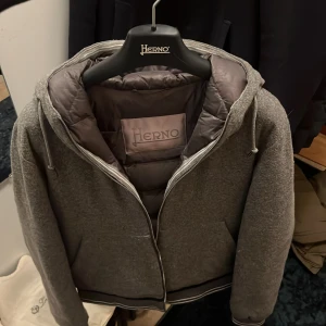 Herno jacka - Säljer en stilren grå jacka från Herno med dragkedja och huva. Jackan har en quiltad insida och ribbade muddar vid ärmslut och nederkant. Perfekt för kyligare dagar.