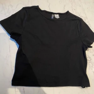 Svart kortärmad topp från Divided - Säljer en enkel och stilren svart kortärmad topp från Divided. Perfekt för en avslappnad look. Toppen har en rund halsringning och är tillverkad i ett mjukt material.