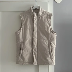 Beige dunväst - Snygg beige dunväst med dragkedja framtill. Perfekt för lager-på-lager under kyliga dagar. Västen har en stilren design med två fickor på sidorna. Hör av er vid intresse! Pris går att diskutera! 