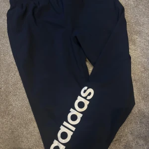 Svarta träningsbyxor från Adidas - Snygga svarta träningsbyxor från Adidas med stort logotryck längs benet. Perfekta för träning eller en avslappnad dag. Byxorna har en elastisk midja för extra komfort.