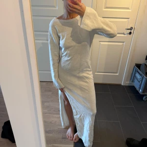 Beige stickad långklänning - Säljer en mysig vit glittrig stickad långklänning med lång ärm och en stilren slits på sidan. Perfekt för kyligare dagar när du vill vara både bekväm och snygg. Klänningen har en rund halsringning och en mjuk, skön passform.