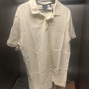 Vit pikétröja från GANT - Säljer en klassisk vit pikétröja från GANT med korta ärmar och krage. Tröjan har en liten broderad logga på bröstet och knappar vid halsen. Perfekt för en stilren look. Dock en liten fläck som garanterat går bort med en glugg tvätt