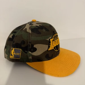 Kamouflage keps med Lakers-logga - Snygg keps med kamouflagemönster och broderad Los Angeles Lakers-logga i gult. Kepsen har en gul skärm och justerbar baksida för perfekt passform. Perfekt för basketfans som vill sticka ut!