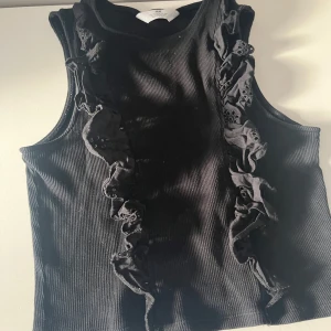 Svart ribbad linnetopp från H&M - Snygg svart ribbad linnetopp från H&M med volangdetaljer framtill. Perfekt för en stilren look. Toppen är ärmlös och har en bekväm passform.