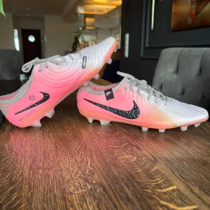 Nike fotbollsskor i vitt och rosa - Snygga fotbollsskor från Nike i en fräsch kombination av vitt och rosa. Skorna har snörning och en svart Nike-logga på sidan. Perfekta för fotbollsplanen med sin stilrena design och funktionalitet.