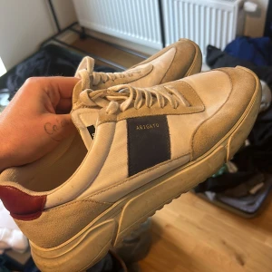 Beige sneakers från Arigato - Säljer ett par snygga beige sneakers från Arigato. De har en stilren design med blå och röda detaljer. Skorna är i bra skick och passar perfekt för vardagsbruk. De har snörning och en bekväm sula. Nypris är ca 2000kr!