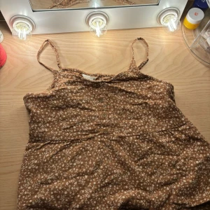 Brun blommig linnetopp från H&M🤎 - Säljer en söt brun linnetopp från H&M med vita blommor. Toppen har smala axelband och knappar framtill. Perfekt för sommardagar eller en avslappnad vårlook. Den är i bra skick och redo för en ny ägare!