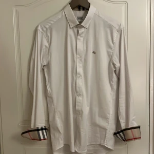 Burberry skjorta - Säljer en stilren vit skjorta från Burberry i storlek M som knappt är använd. Skjortan har långa ärmar och klassiska knappar framtill. Den är broderad med Burberrys ikoniska logga på bröstet och har ett snyggt rutigt mönster på insidan av ärmsluten. Perfekt för både formella och avslappnade tillfällen!🍾 Kom pm vid minsta fundering 🍾🥃