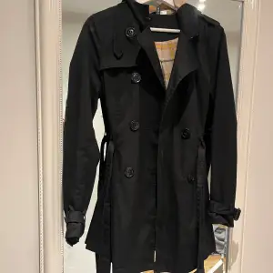 Snygg svart trenchcoat med klassiskt rutigt foder. Jackan har långa ärmar och knappar framtill samt ett bälte i midjan för en elegant look. 